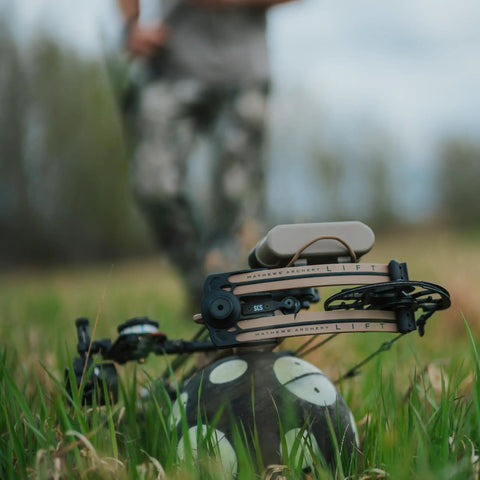 Ultra Portable 9 Inch Sand Filled Archery Target With Carry Handle For On The Go Practice Self Balancing Core Keeps Target Upright After Every Shot Broadhead And Field Point Compatible Great For Backcountry Or Backyard Practice Made In USA in grassy field with bow in foreground