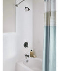 Colony Pro Tub And Shower Trim Kit with bronze finish installed in a white tiled bathroom, showing showerhead and tub faucet.