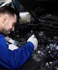 Mechanic in blue uniform inspecting car engine in a garage setting with open hood and engine components visible.