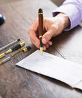 Person writing with 12 Pack Woodturning Pen Kit With Twist Ballpoint Refills Copper Turning Supplies on wooden desk