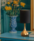 Modern Table Lamp With Brass Droplet Base And Black Linen Drum Shade on a blue table next to a vase of flowers