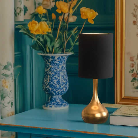Modern Table Lamp With Brass Droplet Base And Black Linen Drum Shade on a blue table next to a vase of flowers
