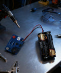 Hands On Soldering Practice Kit For Electronics Newbies with circuit board, batteries, and tools on a workbench.