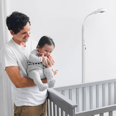 Man holding baby near crib with Floor Stand For Nanit Pro Baby Monitor With Gooseneck Swivel Holder visible in background.