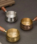 Three stainless steel espresso measuring cups with wooden handles, featuring V-shaped spouts, shown in gold, silver, and bronze.