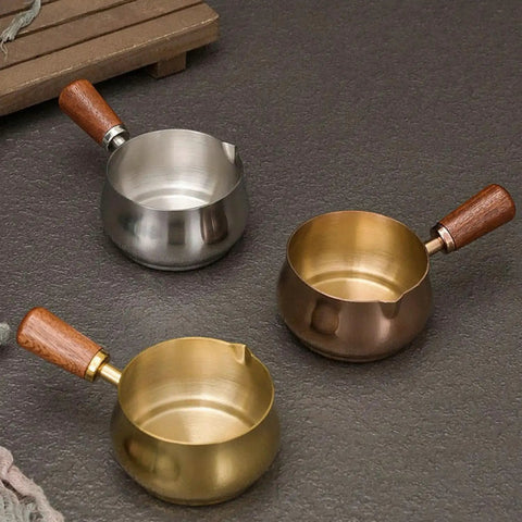 Three stainless steel espresso measuring cups with wooden handles, featuring V-shaped spouts, shown in gold, silver, and bronze.