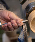 Wood turning chisel with wooden handle held against a rotating wooden spindle on a lathe.