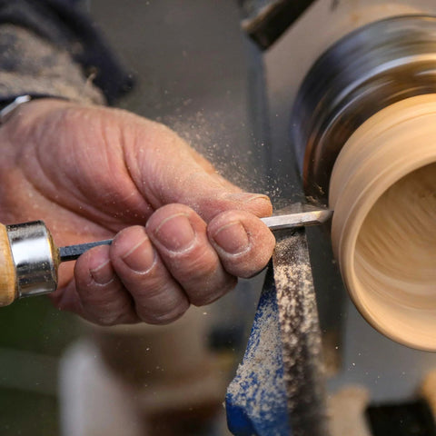 Wood turning chisel with wooden handle held against a rotating wooden spindle on a lathe.
