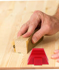 Close-up of a hand using a wooden sanding block on wood, with a red plastic wedge visible on the table.