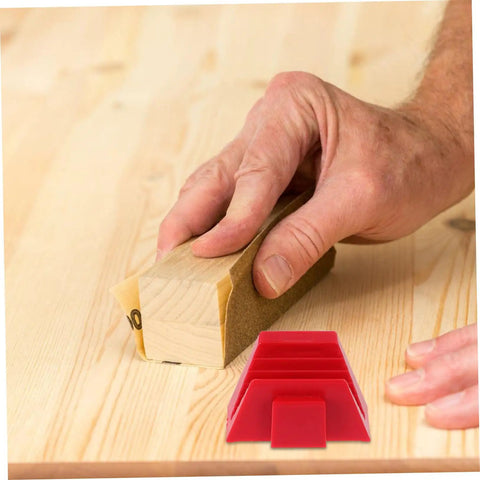 Close-up of a hand using a wooden sanding block on wood, with a red plastic wedge visible on the table.
