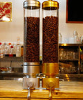 Wall Mounted Transparent Coffee Canister With Gold Trim And Smooth Grip Handle Straight Up Fantastic Dry Food Dispenser For Sugar Rice Nuts Cereals Pet Treats Zero Stress Counter Organizer To Keep Your Pantry Legit with coffee beans inside.