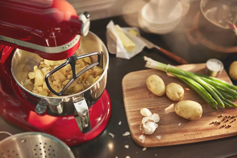 Red KitchenAid mixer with stainless steel paddle attachment mixing potatoes on countertop with ingredients beside it