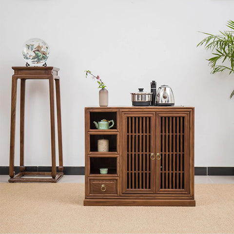 Wooden cabinet with slatted doors and shelves, metal handles, topped with teapots, next to a plant and pedestal with vase