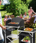 Folding Camping Kitchen Table With Windshield And Storage Cabinet in a backyard setting with people dining, visible fresh produce and grill.