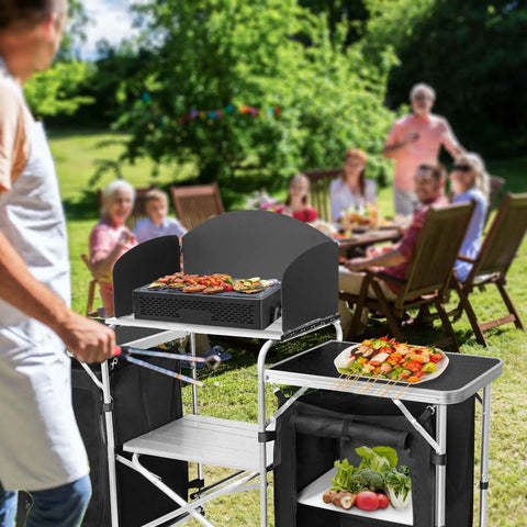 Folding Camping Kitchen Table With Windshield And Storage Cabinet in a backyard setting with people dining, visible fresh produce and grill.