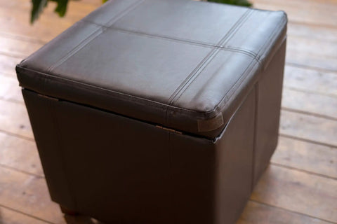 Square brown leather ottoman with stitching details on a wooden floor, closed lid