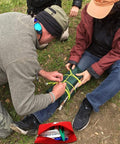 Red first aid kit bag open outdoors; man tying neon yellow cord on a green leg brace while a seated woman helps