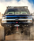 Off-road truck driving through dust with visible front bumper and grille design against a blue sky background.