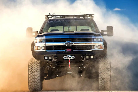 Off-road truck driving through dust with visible front bumper and grille design against a blue sky background.