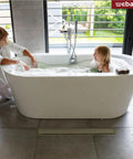 Bathroom with 36 Inch Linear Shower Drain With No-Nonsense Reversible 2-In-1 Cover Tile Insert And Removable Grate In Brushed Gold SUS304 Stainless CUPC Certified Easy Clean Ultra Efficient Drainage For Quick Water Removal In Modern Baths, woman and child in white bathtub, large windows with greenery outside.
