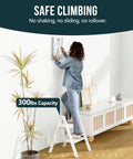 Sturdy 2 Step Ladder With 17 Inch Top Step, 300 Lb Capacity, White Finish, Wider 7.9 Inch Treads For Comfort And Stability, Great For Home, Garage, And DIY Tasks. woman standing on white 2-step ladder hanging wall art indoors