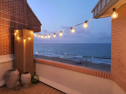 Two Pack 100 Foot Outdoor String Lights With Dimmable G40 LED Bulbs That Are Shatterproof And Weatherproof End To End Connectable Up To Nine Times 100 Feet on a seaside patio at dusk