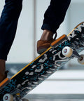 Person performing skateboard trick with patterned deck, view of feet in brown shoes, soft focus background.