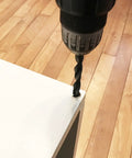 Drill creating hole in white wood surface on hardwood floor, preparing for Brown Wood Wheel Stem Casters Set of Two installation.