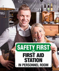 Two people holding a durable aluminum first aid station sign, 8 by 12 inches, for office, shop, factory, warehouse, and public areas.