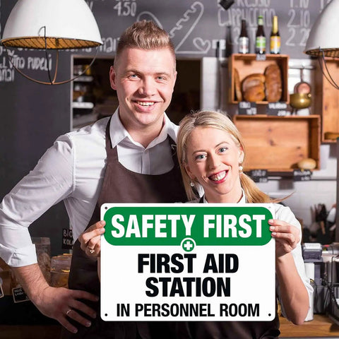 Two people holding a durable aluminum first aid station sign, 8 by 12 inches, for office, shop, factory, warehouse, and public areas.