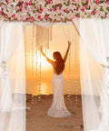 Woman in white dress standing on beach at sunset under floral arch with chiffon curtains.