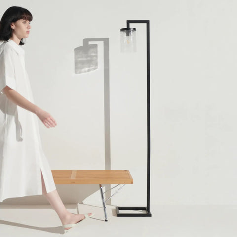 Woman in white dress walking past a minimalist black floor lamp with wooden bench on a white background