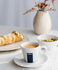 Coffee cup on saucer next to croissant and bowl of nuts on white table with vase and muted decor.