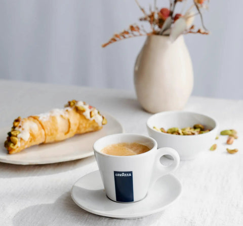 Coffee cup on saucer next to croissant and bowl of nuts on white table with vase and muted decor.