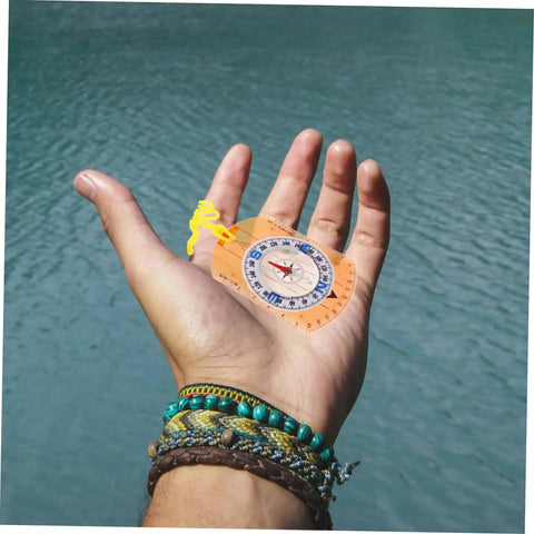 Orienteering Compass Pointer For Outdoor Navigation And Survival Hike Ready Compact Hiking Tool displayed on hand by a lake.