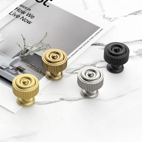 Assorted brushed gold, silver, and black ribbed cabinet knobs displayed on marble surface with magazine decor.