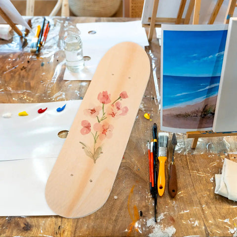 Blank Unfinished 7 Ply Wooden Skateboard Deck With Double Tail For Art Painting And Do It Yourself DIY Custom Builds Replacement Deck For Home Decor And Creative Projects on table with painting tools and floral art.