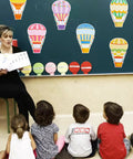60 Colorful Hot Air Balloon Paper Cutouts With 120 Glue Dots, 6 Styles displayed on a classroom blackboard with children seated below.