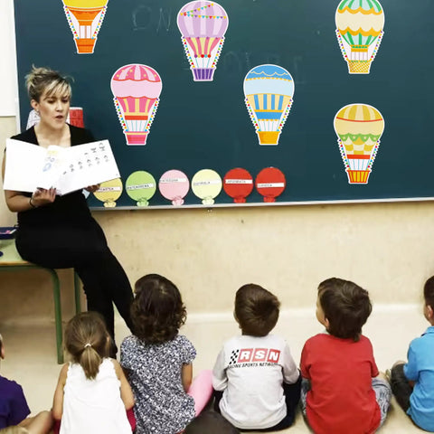 60 Colorful Hot Air Balloon Paper Cutouts With 120 Glue Dots, 6 Styles displayed on a classroom blackboard with children seated below.