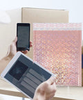 Person using mobile devices next to Rose Pink Holographic Padded Mailers 25x30cm Self Seal Waterproof Shipping Bags 10 Pack for mailing
