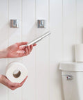 Spring Loaded Toilet Paper Roll Holder Replacement in chrome finish being installed in a bathroom with white walls and stacked toilet paper rolls.