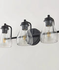 Black Three Light Vanity Light Fixture With Clear Glass Shades For Bathroom Over Mirror, shown mounted on a white wall.