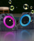 Two Pack Solar Powered Rock Speakers With Lit RGB Lights IP44 Water Resistant Outdoor Speakers For Patio Pool Party Yard Garden USB Rechargeable And USB Charging shown in stone design, each with glowing RGB lights in a garden setting.