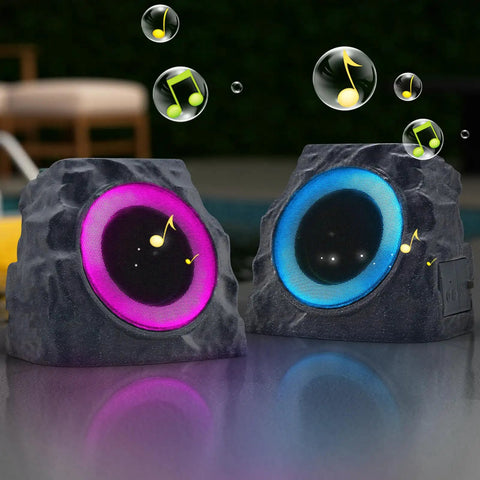 Two Pack Solar Powered Rock Speakers With Lit RGB Lights IP44 Water Resistant Outdoor Speakers For Patio Pool Party Yard Garden USB Rechargeable And USB Charging shown in stone design, each with glowing RGB lights in a garden setting.