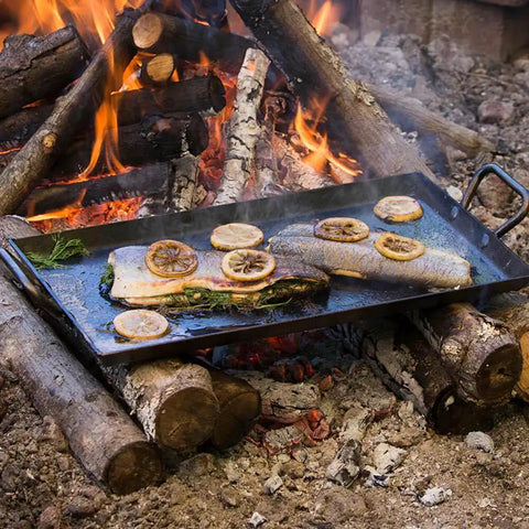 Cooking fish with lemons on a seasoned carbon steel griddle with dual-handle flat top over campfire, 18 by 10 inches.