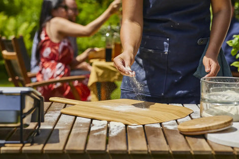 Person sprinkling flour on a Heavy Duty 14 Inch Bamboo Pizza Peel And Wooden Spatula Paddle For Backyard Cookouts.