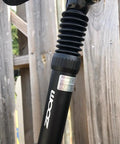 Close-up of a bike suspension seat post with adjustable spring coil damper, black, visible in a wooden background.