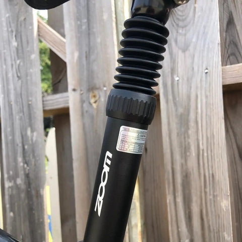 Close-up of a bike suspension seat post with adjustable spring coil damper, black, visible in a wooden background.
