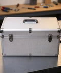Heavy Duty Silver Metal Tool Box With Reinforced Corners Secure Carry Handle Tough As Nails For Workshop And Field Repair Jobs Keeps All Gear Locked And Loaded seen from the front on a workshop table.