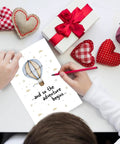 Child's hands holding Hot Air Balloon Adventure Baby Shower Card For New Parents Funny Congratulations Card Blank Inside 8x5.3 Inch Size With Envelope Perfect For Pregnant Couples And Newborn Celebrations surrounded by hearts and gifts.